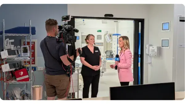 Woman in pink suit interviewing a woman in hospital scrubs in front of room while camera man stands by recording.
