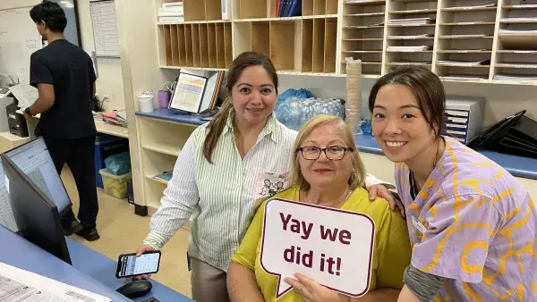 3 women smiling behind a desk with one holding up a sign 'Yay we did it!'.
